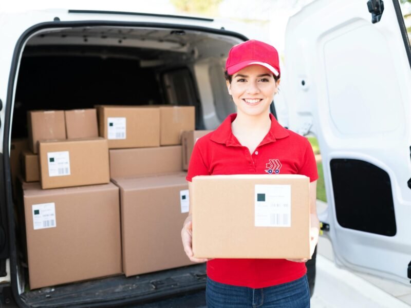 Portrait of a beautiful delivery young woman smiling while holding a package in front of a white van. Female courier working and delivering a parcel