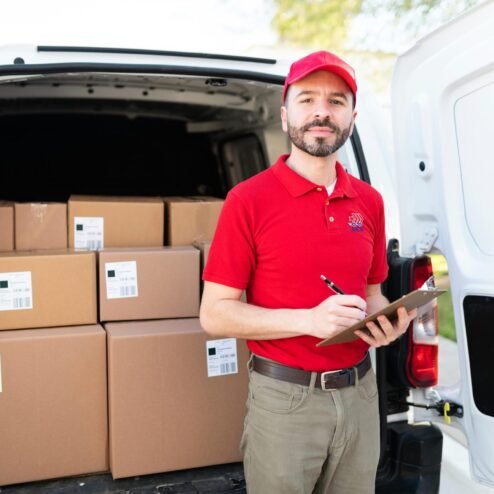 portrait-of-a-male-courier-preparing-to-make-deliv-2025-10-27-09-22-50-utc Handsome latin man wearing a red uniform and standing in the back of a white van. Delivery man making eye contact while holding a clipboard and a pen